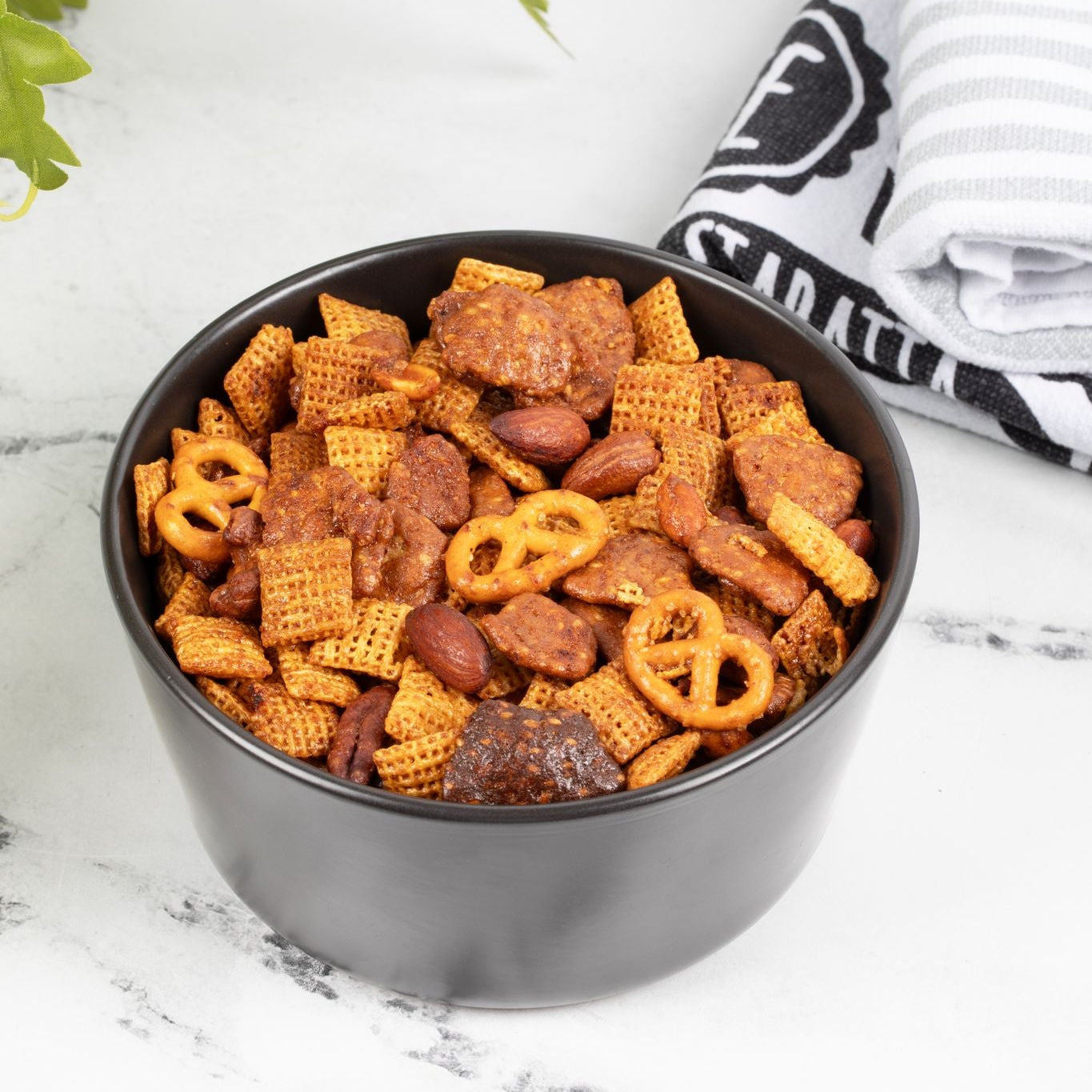 Black bowl of snack mix on a white surface with a plant and towel in the background